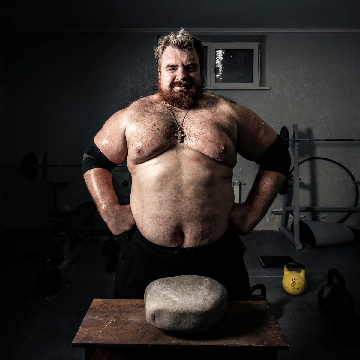 A strongman posing with a training stone and kettlebells, the human scale of what 120 kg of lifting looks like