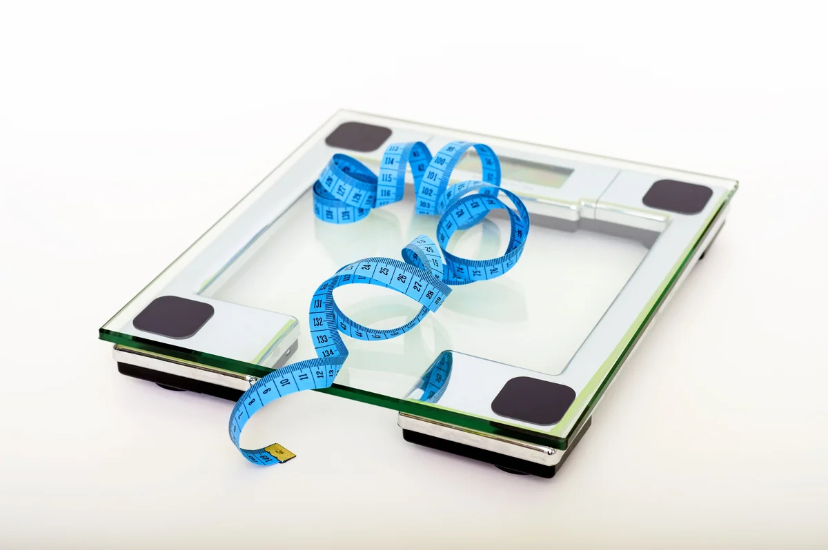 A clear glass digital bathroom scale with a blue measuring tape curled on top, the two tools people use to track 150 lb goals
