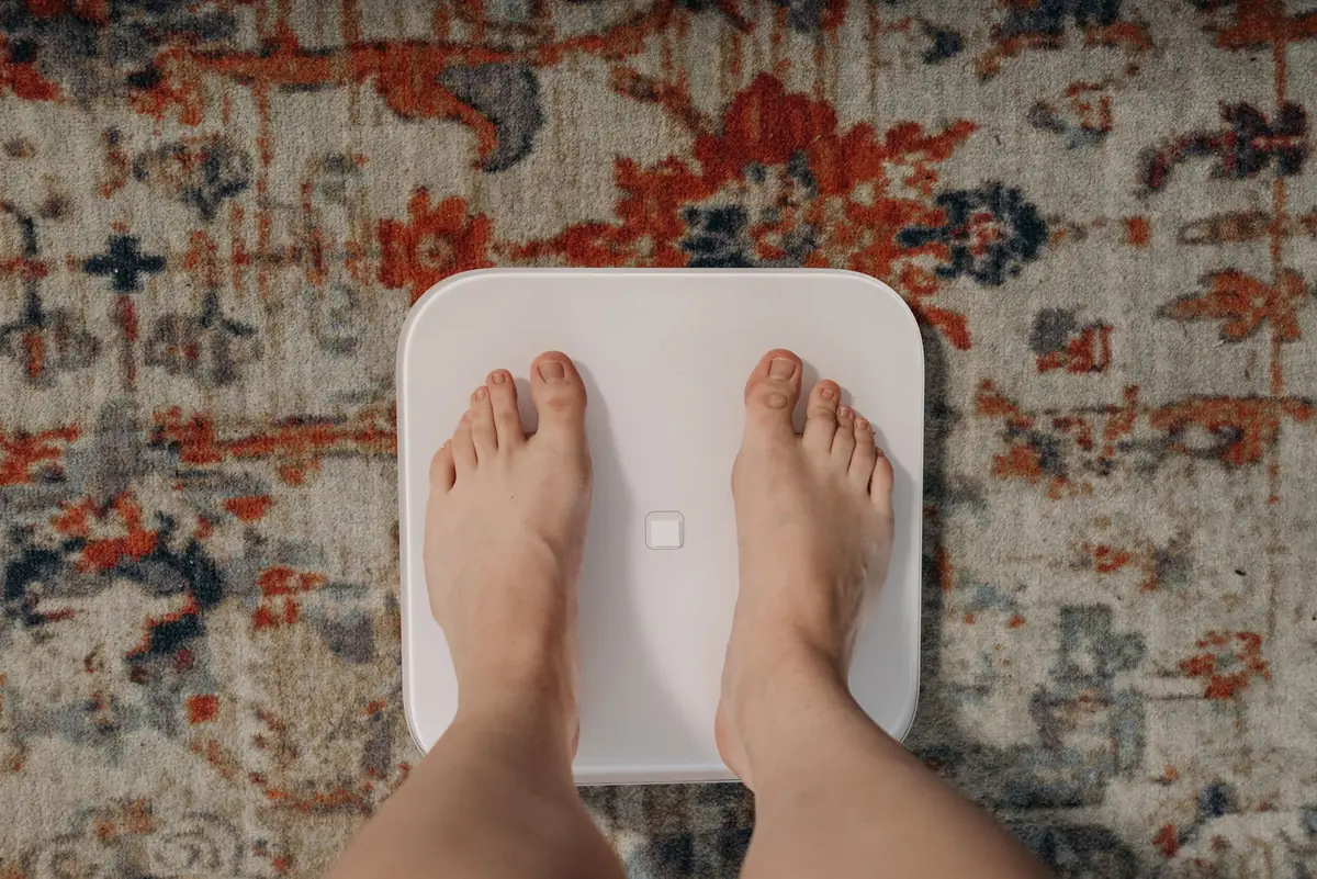 Looking down at bare feet on a white digital bathroom scale on a patterned rug, the familiar home moment before 50 kg turns into stone