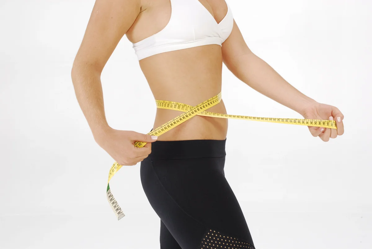 A woman in gym leggings measuring her waist with a yellow tape measure, the honest check behind the 13 stone question