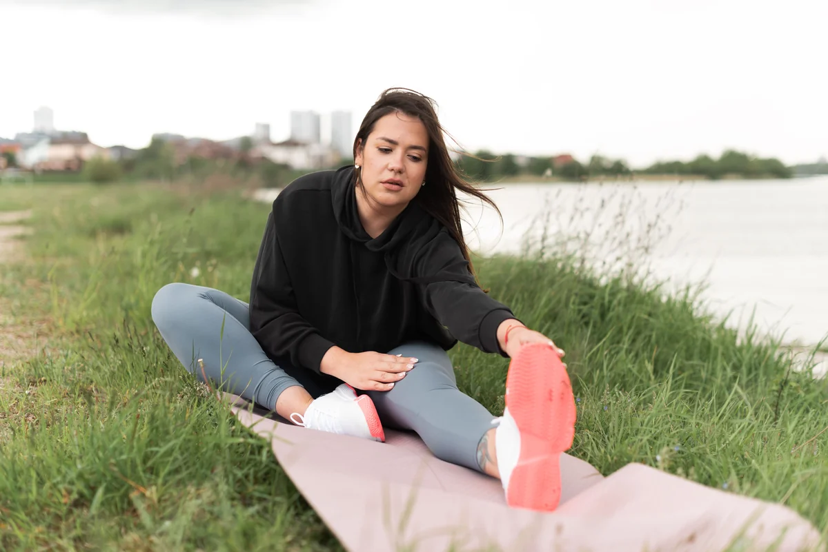 A woman stretching on a yoga mat by a riverside in a city park, a calm start to the 16 stone question