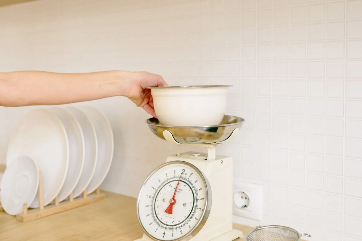 A hand setting a bowl on a vintage kitchen scale, the dial reading pounds before they turn into stone